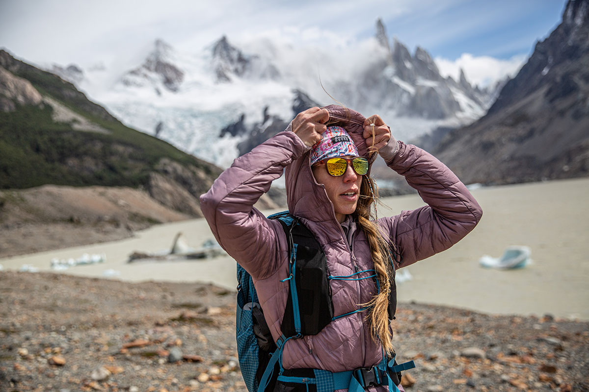 A woman hiking in the snow peaked mountains dons her hood in the wind.